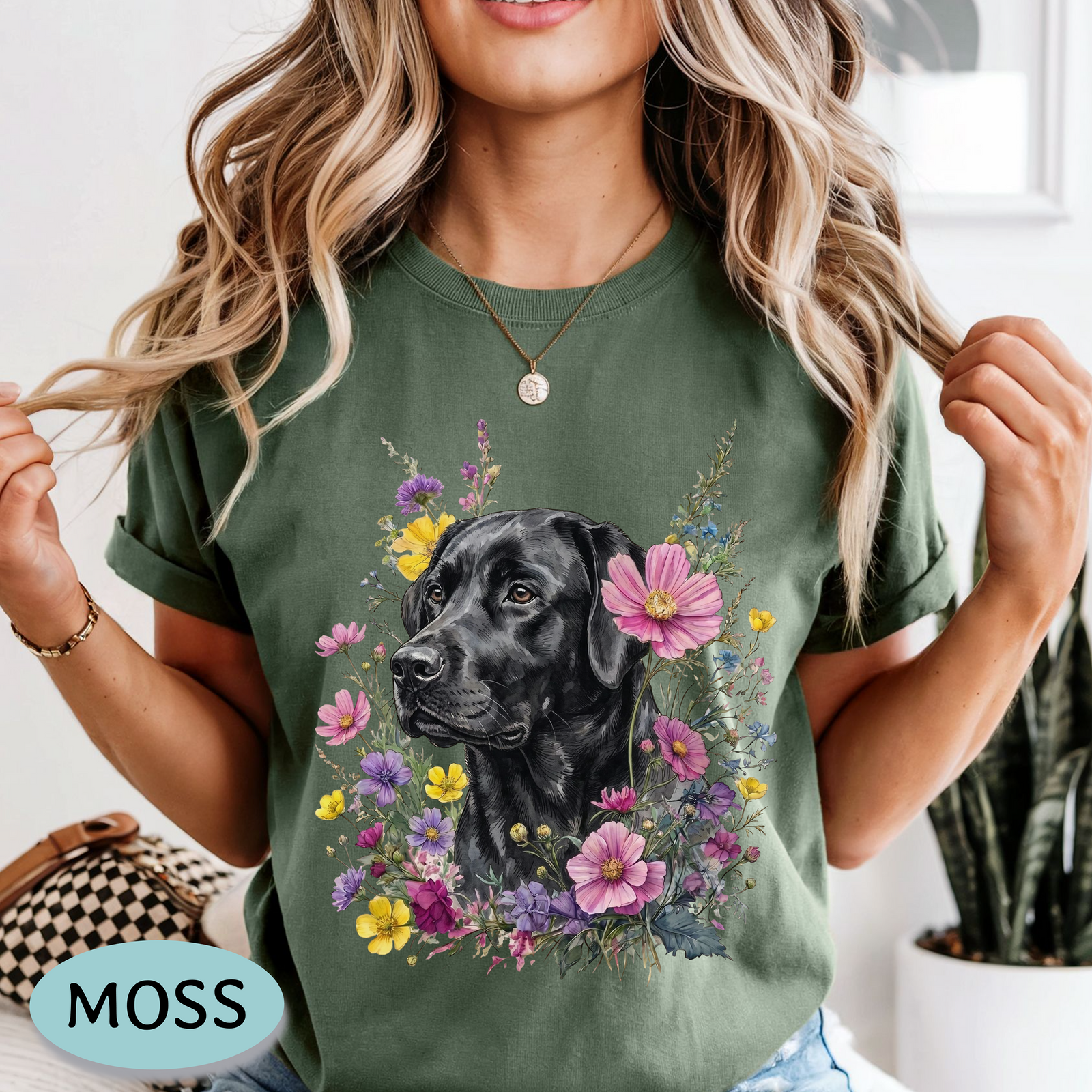 A woman wearing a sage green t-shirt with a graphic of a black Labrador retriever surrounded by pink, purple, and yellow flowers. The image has a 'MOSS' brand label overlay in the bottom left corner.