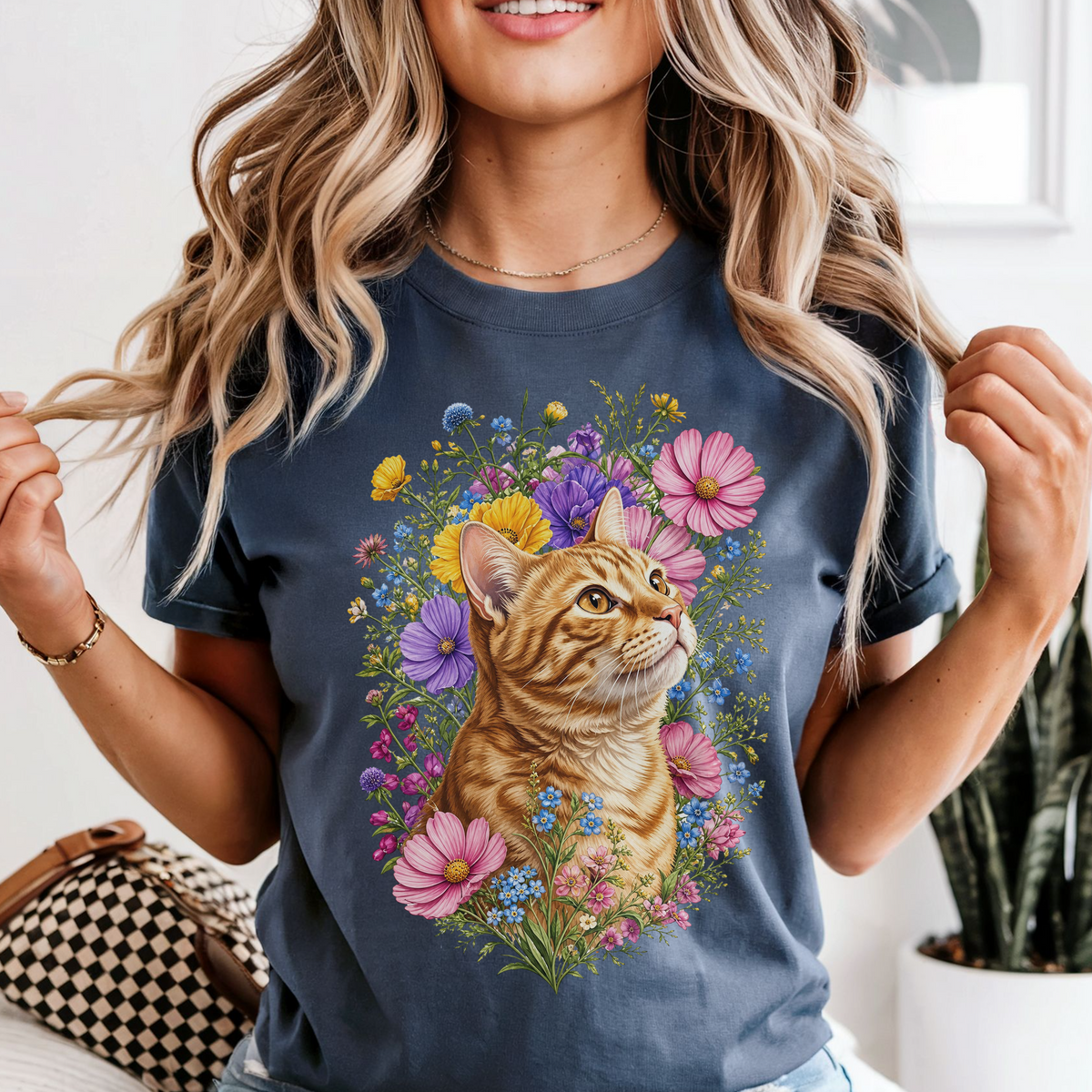 A woman wearing a navy blue graphic t-shirt featuring a detailed illustration of an orange tabby cat surrounded by colorful wildflowers. The photo is taken in a bright indoor setting with a potted plant in the background.