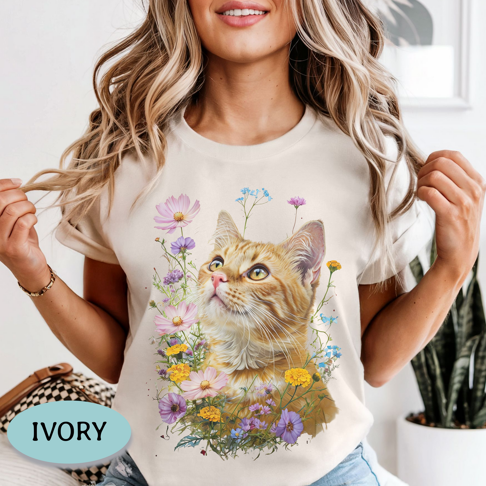 A woman wearing a dark charcoal gray graphic t-shirt featuring a detailed print of an orange tabby cat surrounded by colorful wildflowers. The image is styled as a lifestyle shot with a potted plant in the background and a light blue badge in the bottom-left corner reading 'PEPPER'.