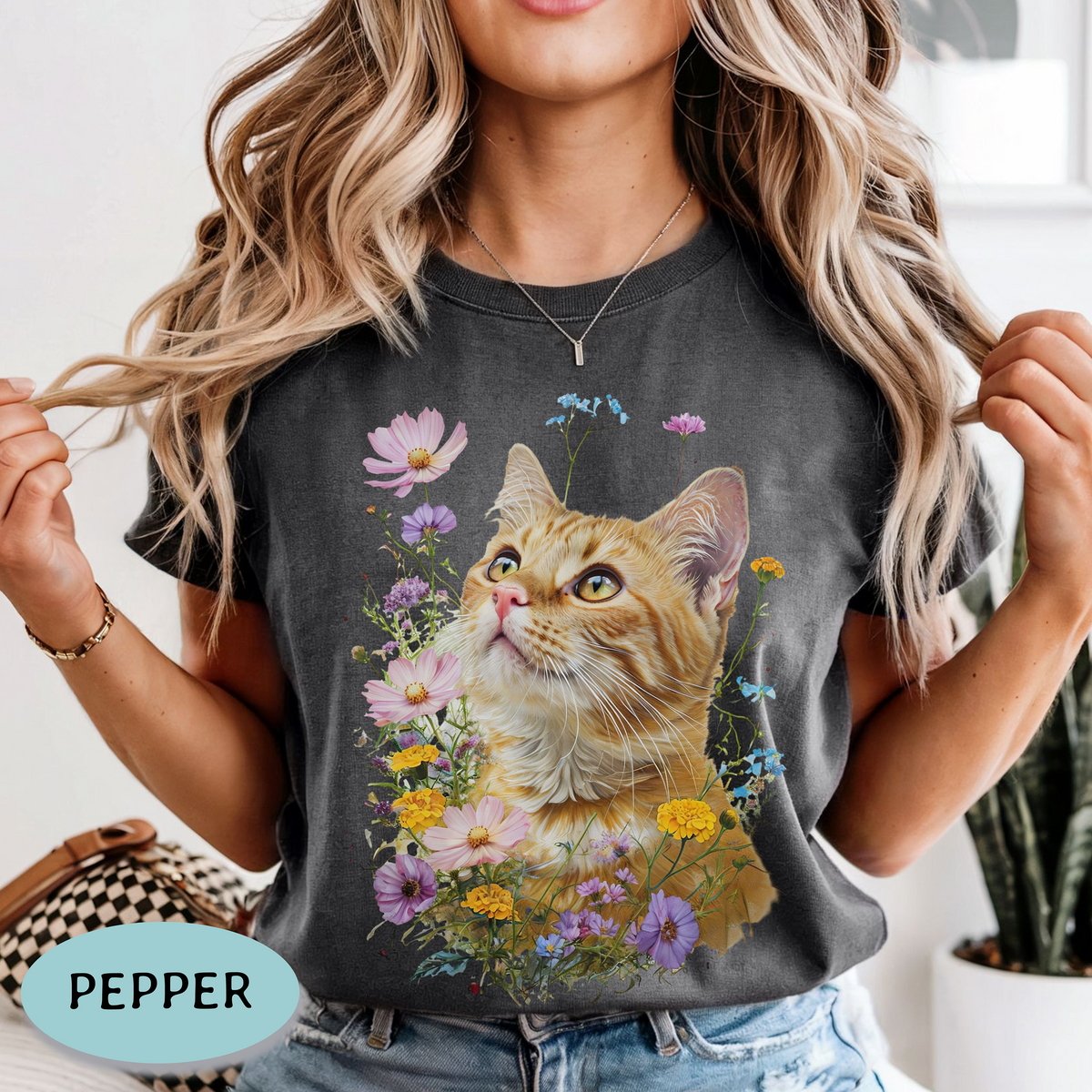 A woman wearing a dark charcoal gray graphic t-shirt featuring a detailed print of an orange tabby cat surrounded by colorful wildflowers. The image is styled as a lifestyle shot with a potted plant in the background and a light blue badge in the bottom-left corner reading 'PEPPER'.