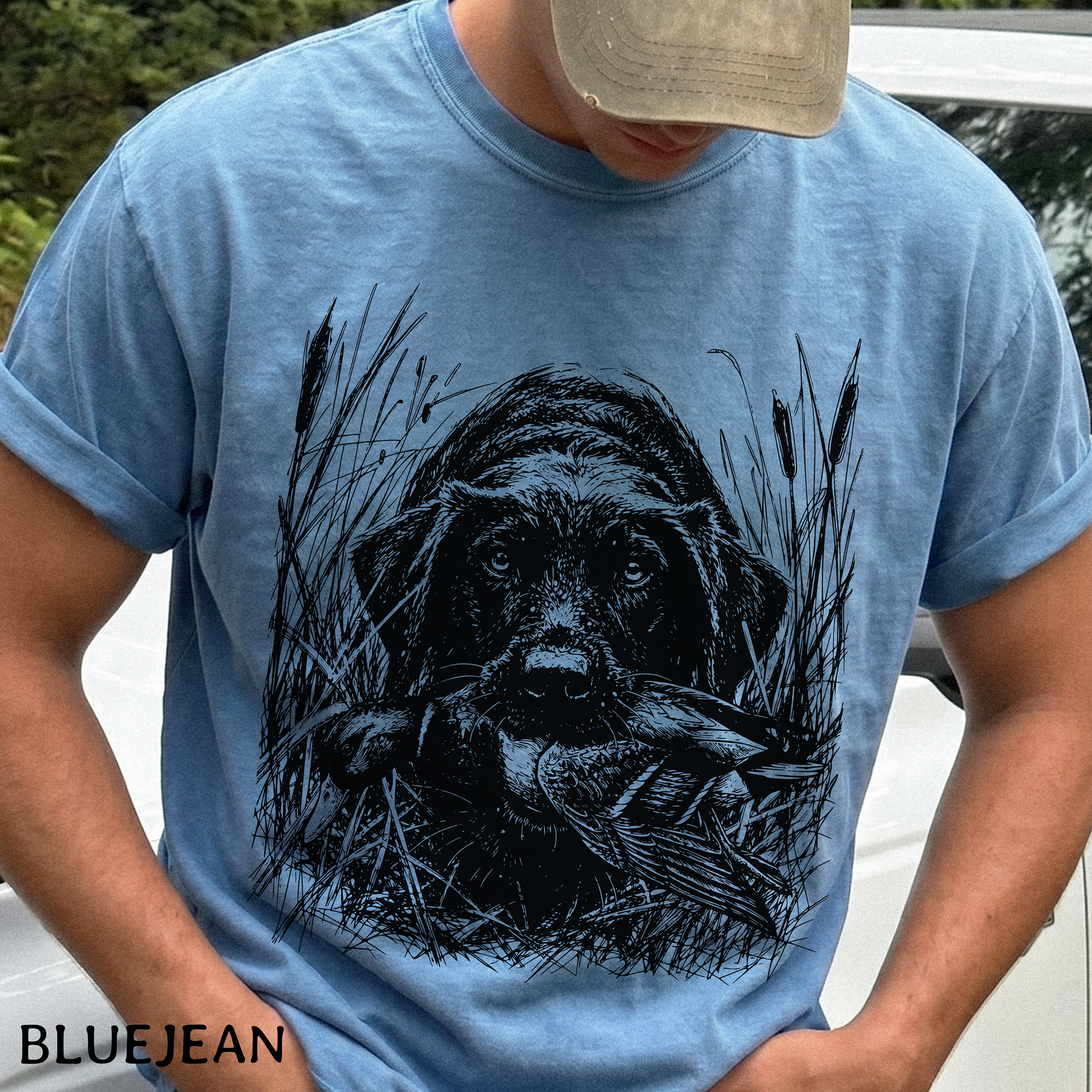 A man wearing a light blue t-shirt with a black graphic print of a dog holding a duck in its mouth, set against a background of reeds. The brand name 'BLUEJEAN' is printed in black text in the bottom-left corner.