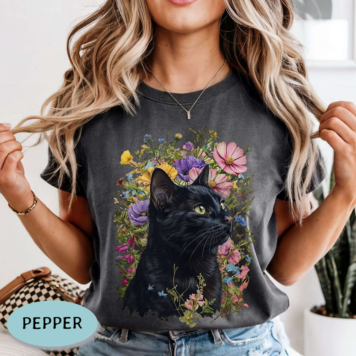 A woman wearing a pepper gray t-shirt with a graphic print of a black cat surrounded by colorful wildflowers. The image is a lifestyle shot with a light-colored indoor background.