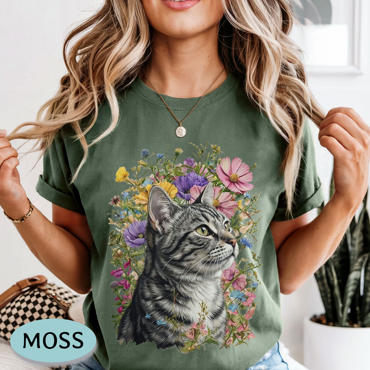 A woman wearing a sage green t-shirt with a graphic print of a gray tabby cat surrounded by colorful wildflowers. The image is a lifestyle shot with a 'MOSS' brand label overlay in the bottom left corner.