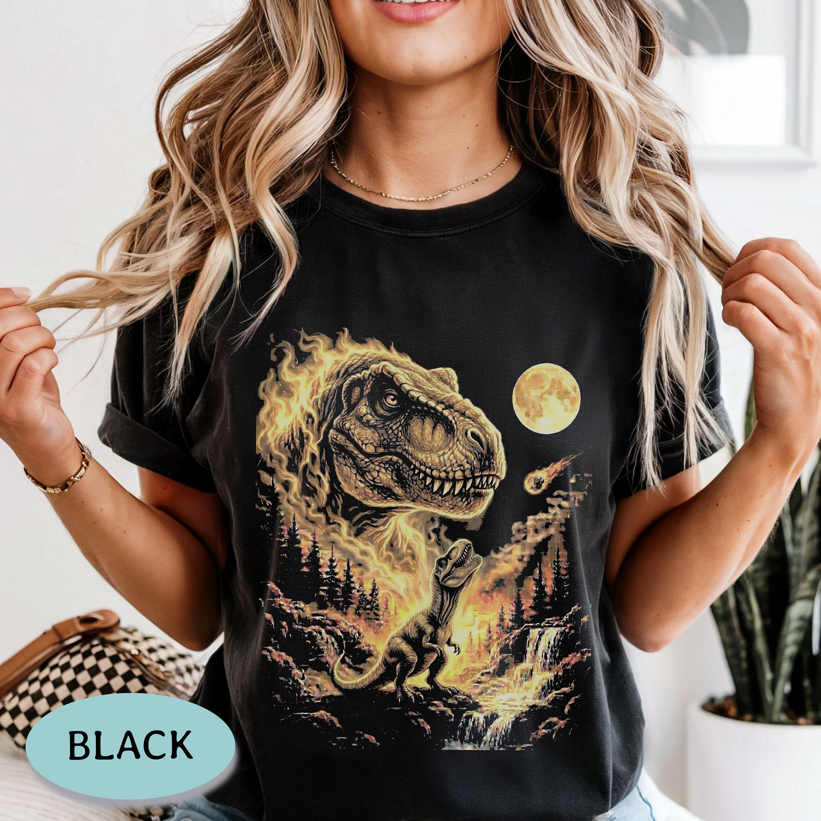 A woman wearing a black graphic T-shirt with a fiery dinosaur scene, holding her hair out of the frame. A circular badge in the lower left says BLACK.