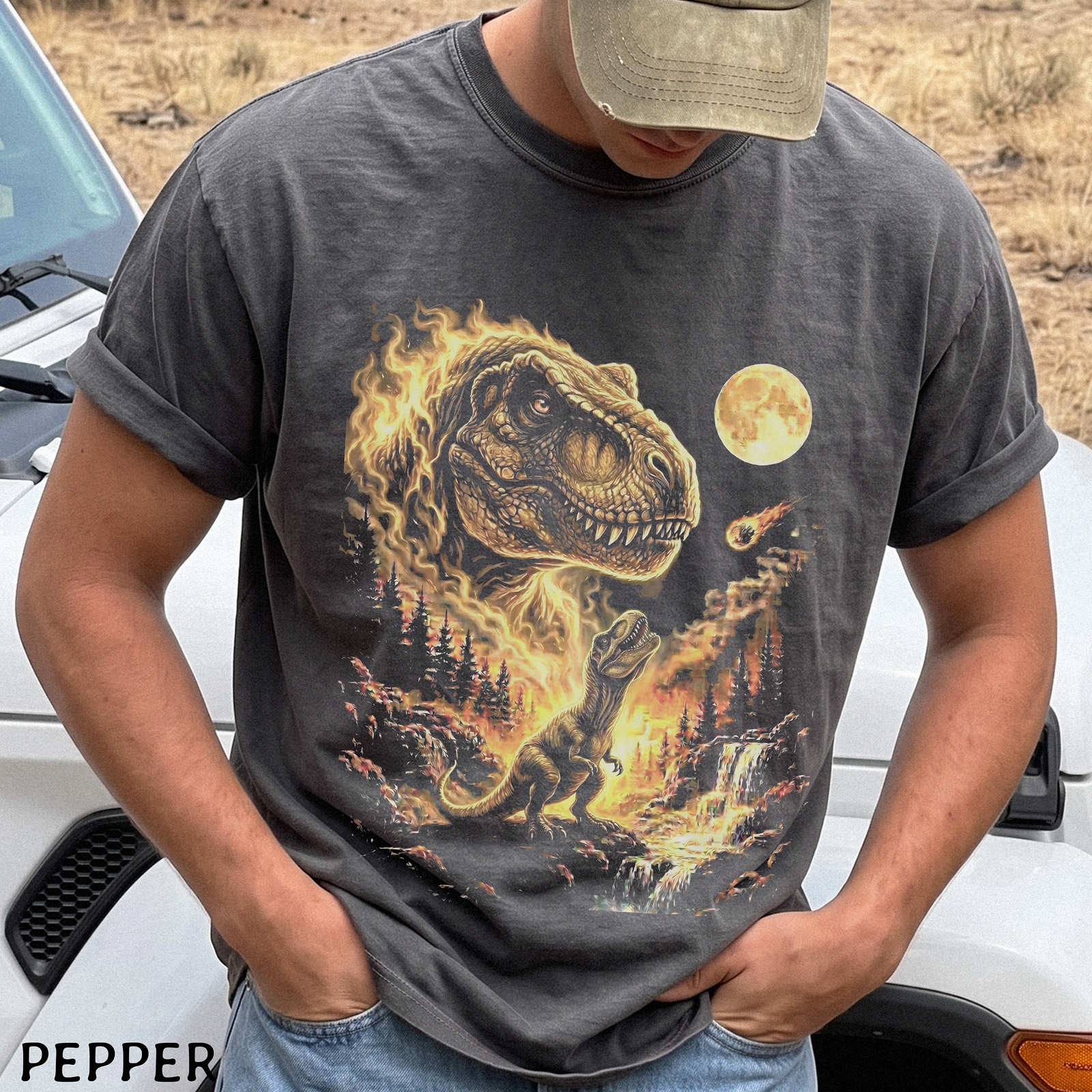A man wearing a dark gray graphic t-shirt with a fiery T-Rex design, standing outdoors in front of a white vehicle. The brand name 'PEPPER' is visible in the bottom-left corner.