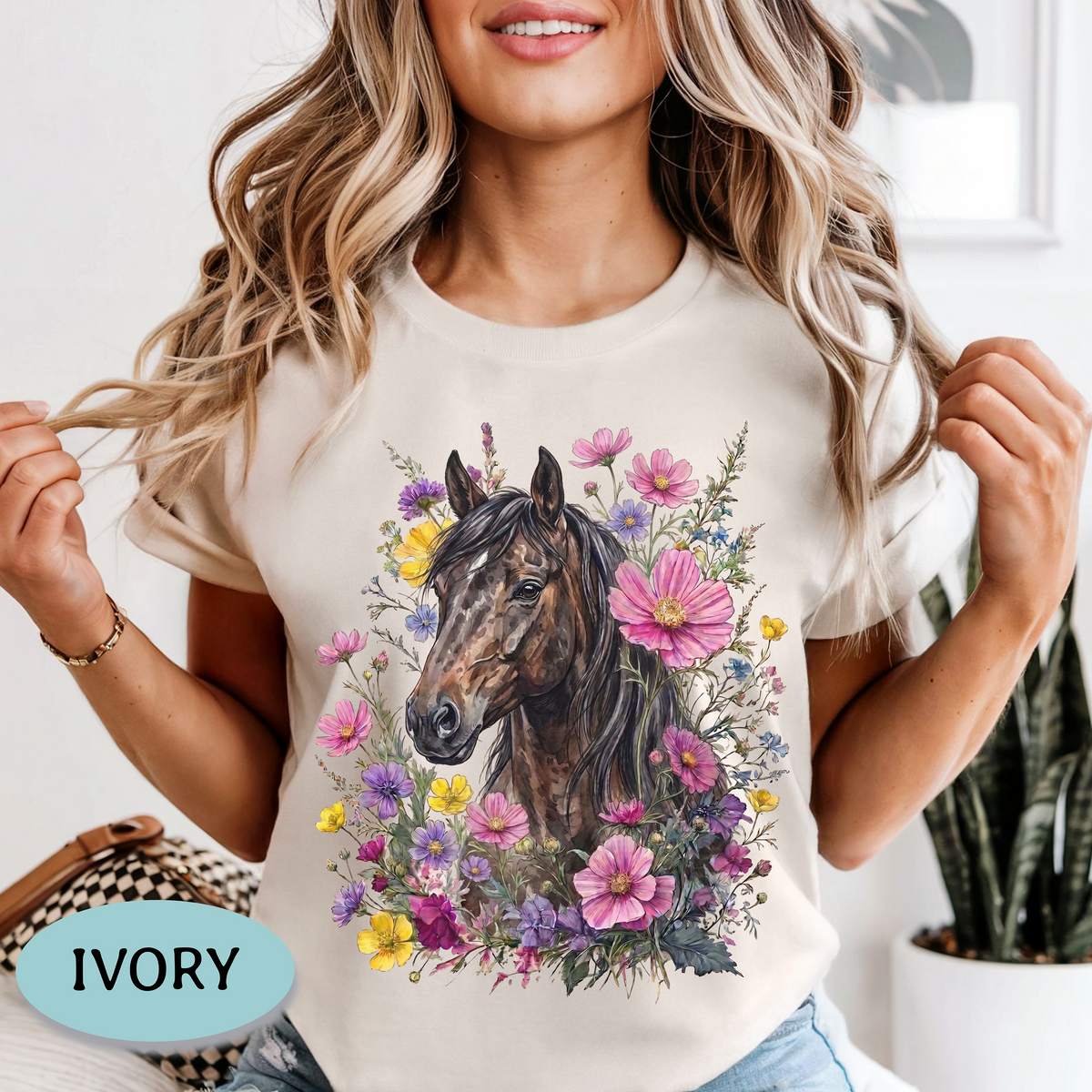 A woman wearing an ivory short-sleeve crewneck t-shirt with a graphic print of a brown horse surrounded by pink, purple, and yellow flowers. A light blue oval label in the bottom left corner reads 'IVORY'.