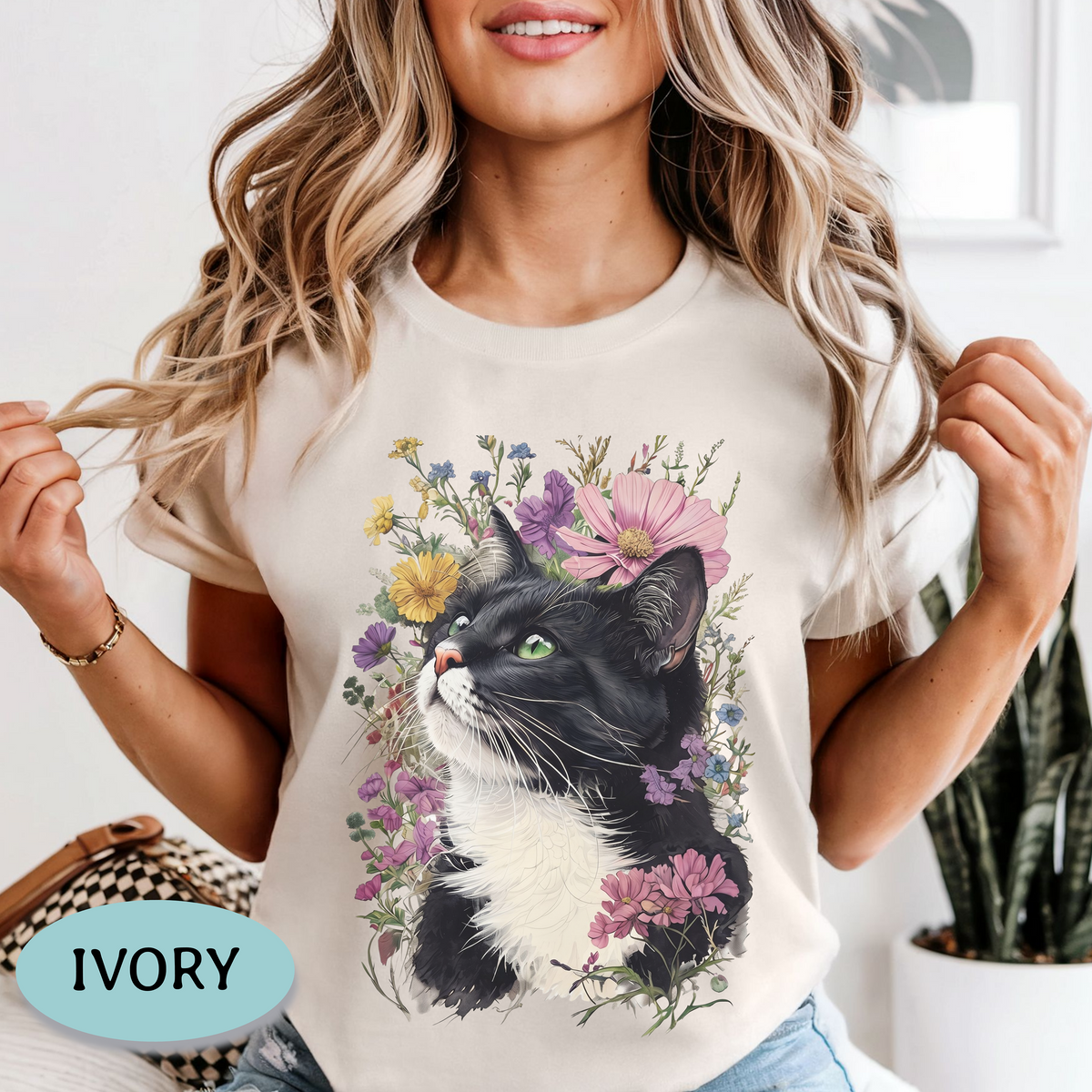 A woman wearing an ivory short-sleeve crewneck t-shirt with a graphic print of a black and white cat surrounded by colorful wildflowers. A light blue oval badge in the bottom-left corner reads 'IVORY'.