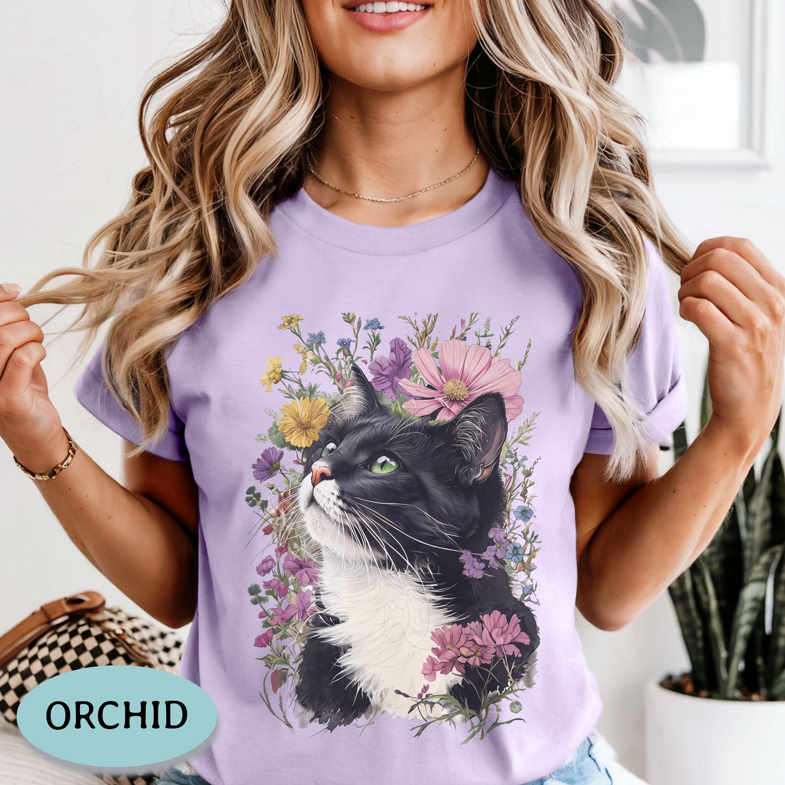 A woman wearing an ivory short-sleeve crewneck t-shirt with a graphic print of a black and white cat surrounded by colorful wildflowers. A light blue oval badge in the bottom-left corner reads 'IVORY'.
